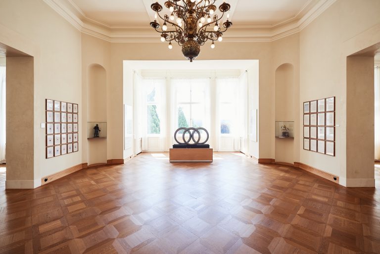 Exhibition room with parquet flooring and a chandelier; in the center stands a sculpture of three interlocking rings, framed pictures hang on the walls.
