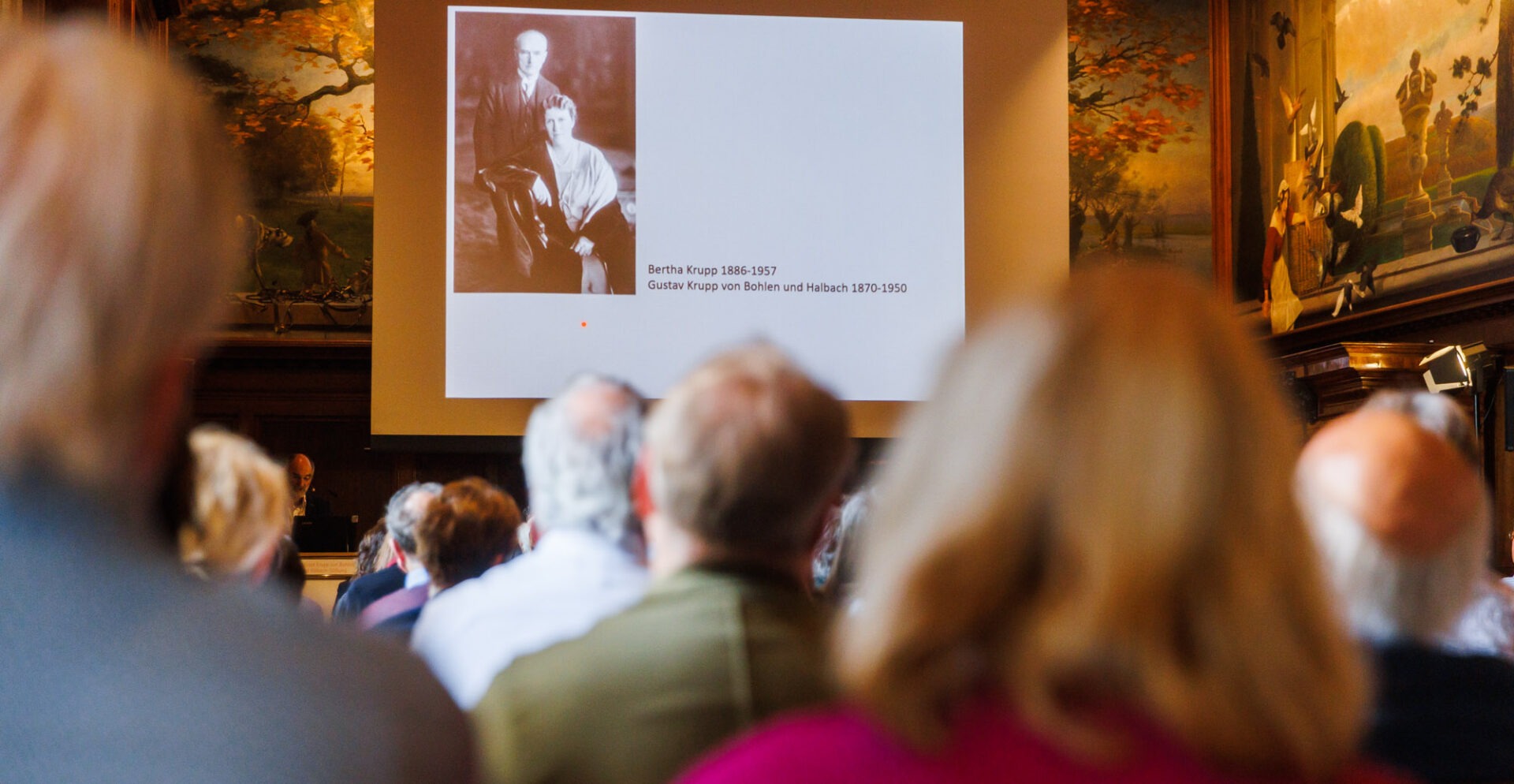 Im Vordergrund sind Besucher eines Vortrags zu sehen, die auf eine Leinwand im Hintergrund schauen mit einem Foto von Bertha Krupp und Gustav Krupp von Bohlen und Halbach schauen.
