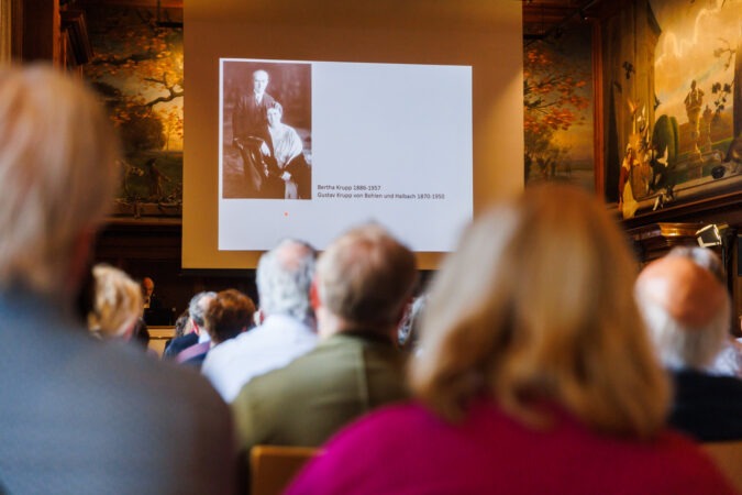 Im Vordergrund sind Besucher eines Vortrags zu sehen, die auf eine Leinwand im Hintergrund schauen mit einem Foto von Bertha Krupp und Gustav Krupp von Bohlen und Halbach schauen.