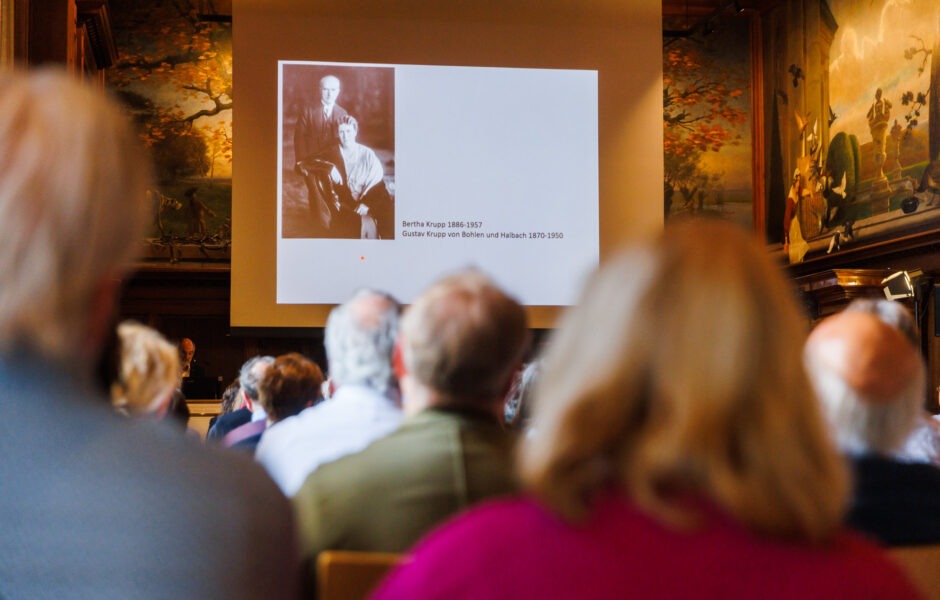Im Vordergrund sind Besucher eines Vortrags zu sehen, die auf eine Leinwand im Hintergrund schauen mit einem Foto von Bertha Krupp und Gustav Krupp von Bohlen und Halbach schauen.