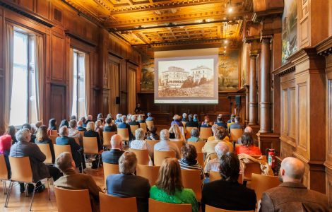 Audience attending a presentation in the dining hall of Villa Hügel, a historic wood-paneled room with an ornate ceiling; a historical photograph of Villa Hügel is displayed on the screen.