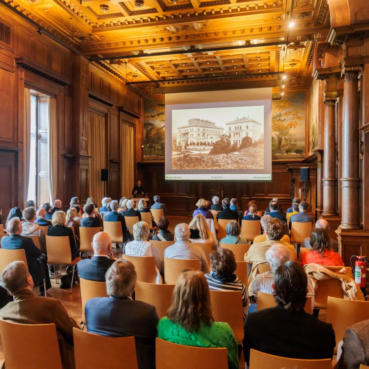 Publikum bei einer Präsentation im Speisesaal der Villa Hügel, einem historischen, holzvertäfelten Saal mit Deckenverzierung; auf der Leinwand ist ein historisches Foto der Villa Hügel zu sehen.