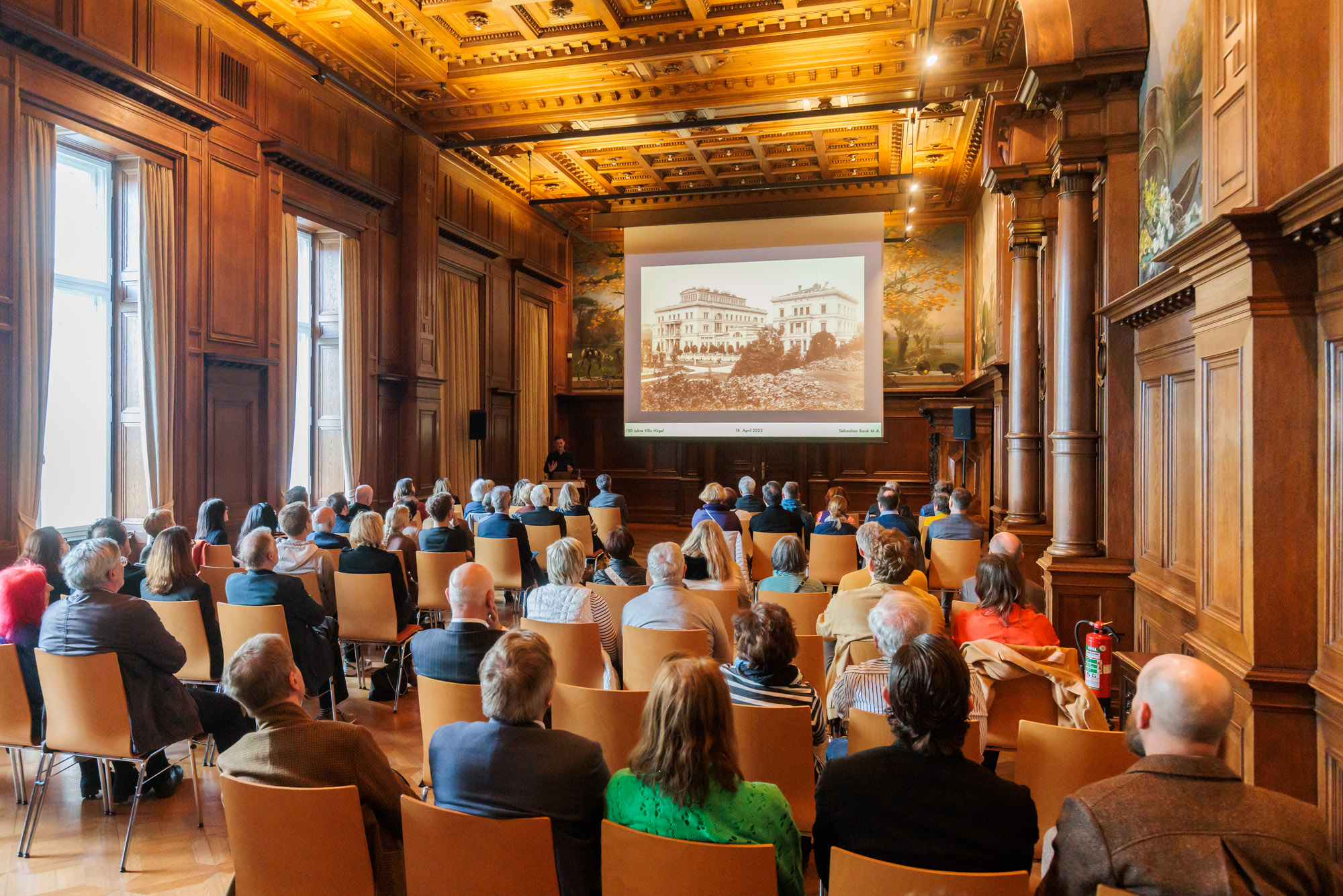 Publikum bei einer Präsentation im Speisesaal der Villa Hügel, einem historischen, holzvertäfelten Saal mit Deckenverzierung; auf der Leinwand ist ein historisches Foto der Villa Hügel zu sehen.