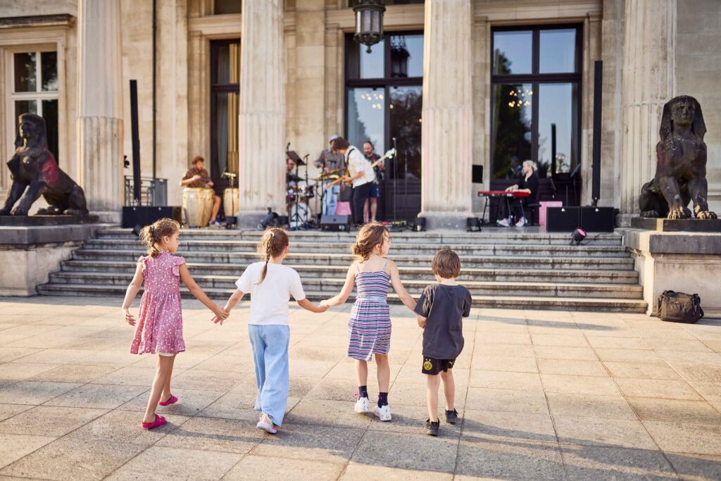 Vier Kinder halten sich an den Händen im Sonnenschein vor der Südfassade der Villa Hügel