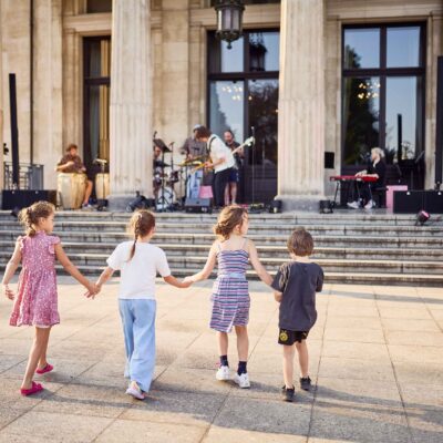 Vier Kinder halten sich an den Händen im Sonnenschein vor der Südfassade der Villa Hügel