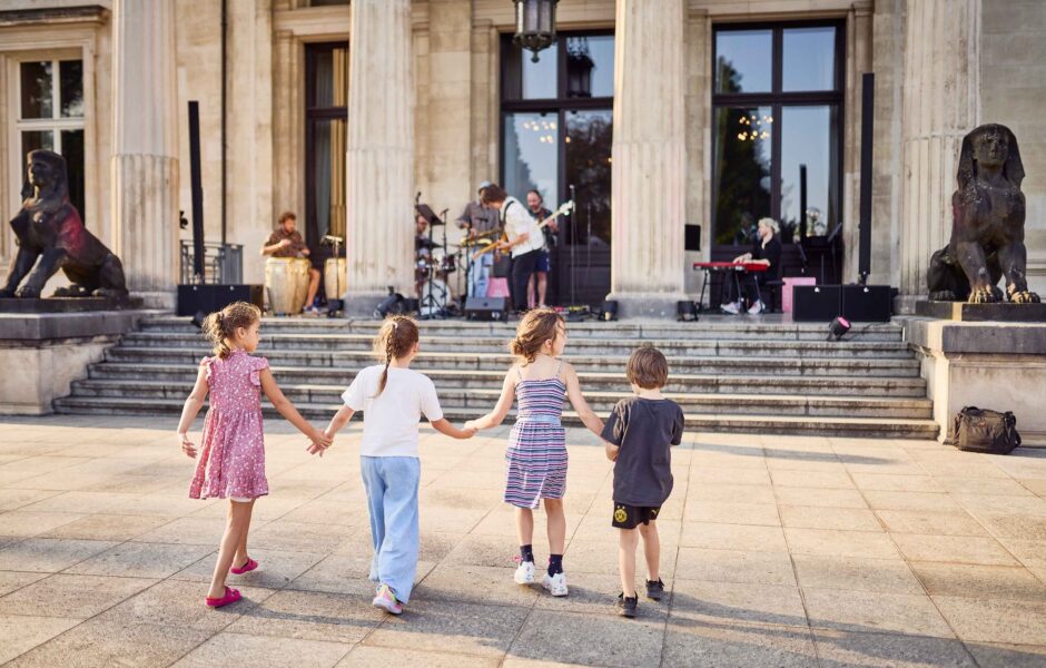 Vier Kinder halten sich an den Händen im Sonnenschein vor der Südfassade der Villa Hügel