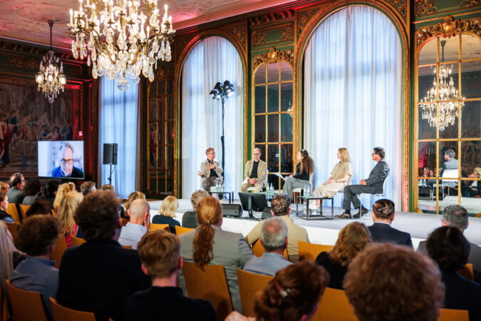 Vier Personen sitzen im Gartensaal der Villa Hügel, einem reich geschmückten und verzierten Raum im LouisIV-Stil, auf einem Podium. Im Vordergrund ist PUblikum zu sehen, links ist ein weiterer Teilnhemer auf einem Fernseher zugeschaltet.