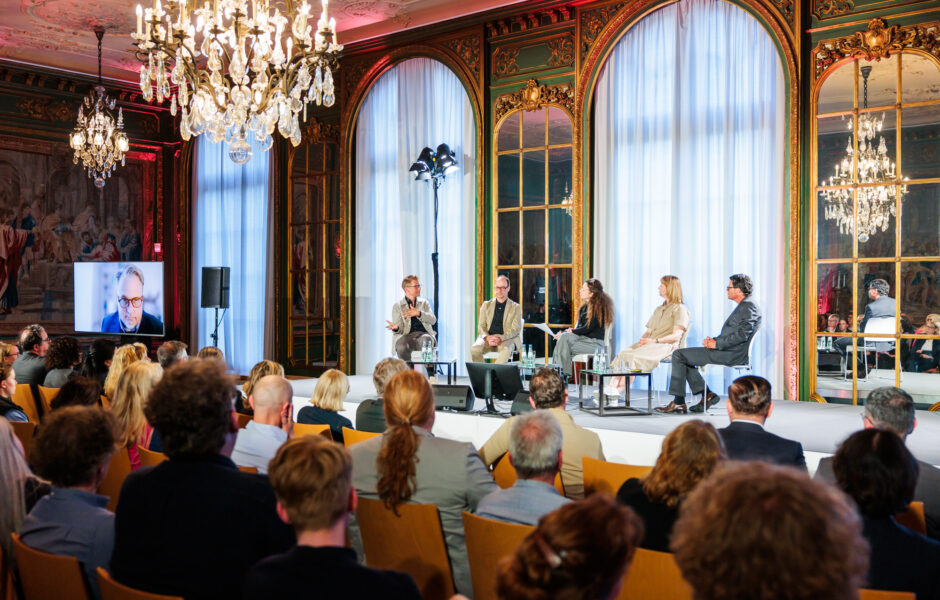 Four people sit on a podium in the Garden Hall of Villa Hügel, a richly decorated and ornate room in the Louis IV style. The audience can be seen in the foreground, with another participant on a television to the left.