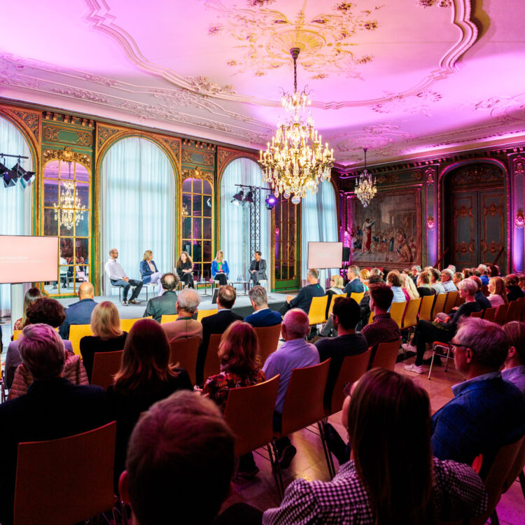 Blick in den Gartensaal, in dem fünf Personen auf der Bühne sitzen vor einem Publikum