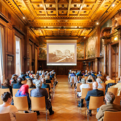 Publikum bei einer Präsentation im Speisesaal der Villa Hügel, einem historischen, holzvertäfelten Saal mit Deckenverzierung; auf der Leinwand ist ein historisches Foto der Villa Hügel zu sehen.