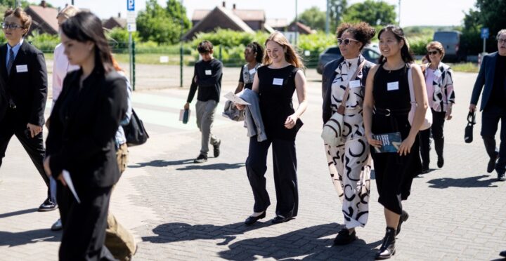 Eine Gruppe von Menschen in formeller Kleidung geht gemeinsam draußen auf einem gepflasterten Weg. Die Sonne scheint, einige Personen lächeln und unterhalten sich. Im Hintergrund sind Wohnhäuser, Bäume und Autos zu sehen.