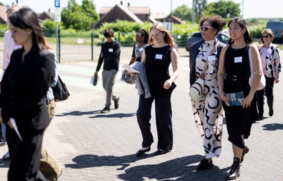 Eine Gruppe von Menschen in formeller Kleidung geht gemeinsam draußen auf einem gepflasterten Weg. Die Sonne scheint, einige Personen lächeln und unterhalten sich. Im Hintergrund sind Wohnhäuser, Bäume und Autos zu sehen.