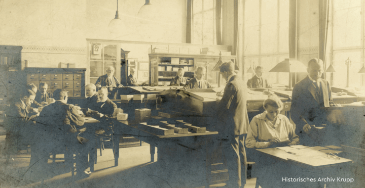 Ein historrisches Foto in Sepia, das Menschen in einem Büro stehend und sitzend an Schreibtischen zeigt.