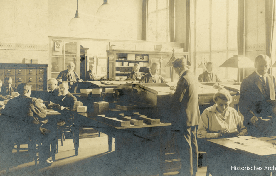 Ein historrisches Foto in Sepia, das Menschen in einem Büro stehend und sitzend an Schreibtischen zeigt.