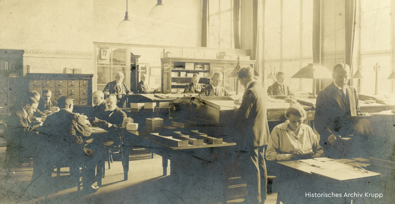 Ein historrisches Foto in Sepia, das Menschen in einem Büro stehend und sitzend an Schreibtischen zeigt.