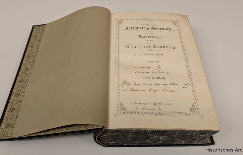 Eine aufgeschlagene Bibel liegt auf einem Tisch. Auf der erste Seite ist zu lesen: "In gesegnetem Gebrauch und zum Andenken an den Tag Ihrer Trauung den 15. Oktober 1906 erhielten von dem Priester aus Werden a.d. Ruhr dieses Bibelbuch: Gustav Georg Friedrich Maria Krupp von Bohlen und Halbach und Bertha Krupp."