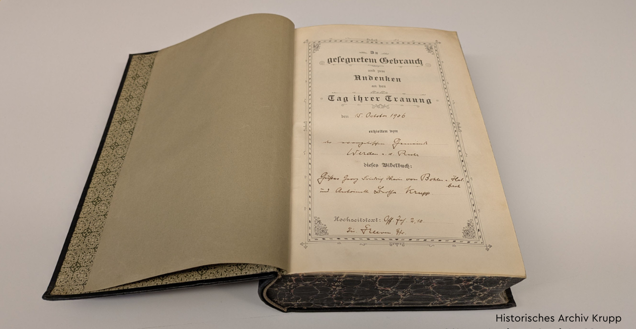 An open Bible lies on a table. The first page reads: ‘In blessed use and in memory of the day of your wedding on 15 October 1906, the priest from Werden a.d. Ruhr gave this Bible book to Gustav Georg Friedrich Maria Krupp von Bohlen und Halbach and Bertha Krupp.’