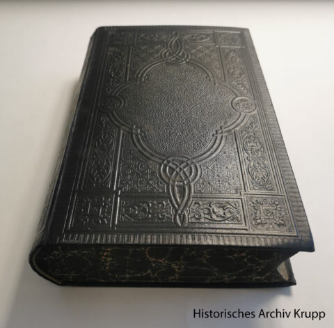 Black embossed Bible with elaborately decorated cover in historical style. The book lies closed on a light-coloured background. The source ‘Historisches Archiv Krupp’ is indicated at the bottom right.