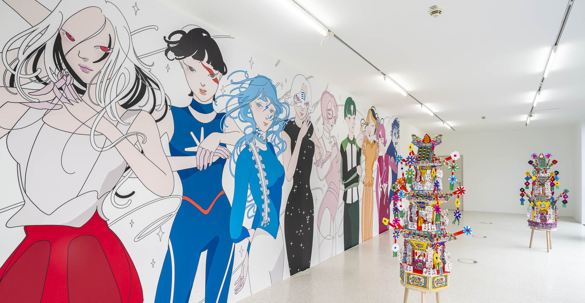 Eine moderne Kunstausstellung in einem hell erleuchteten Galerieraum. An der linken Wand ist ein großes, farbenfrohes Wandgemälde mit stilisierten, anime-inspirierten Figuren in kräftigen Farben zu sehen. Im Raum stehen zwei aufwendig dekorierte, bunte Skulpturen auf Holzstühlen, die aus Papier, Perlen und kleinen Objekten bestehen. Die Szenerie vermittelt eine verspielte und kulturell vielfältige Atmosphäre.