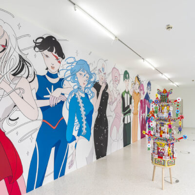 Eine moderne Kunstausstellung in einem hell erleuchteten Galerieraum. An der linken Wand ist ein großes, farbenfrohes Wandgemälde mit stilisierten, anime-inspirierten Figuren in kräftigen Farben zu sehen. Im Raum stehen zwei aufwendig dekorierte, bunte Skulpturen auf Holzstühlen, die aus Papier, Perlen und kleinen Objekten bestehen. Die Szenerie vermittelt eine verspielte und kulturell vielfältige Atmosphäre.