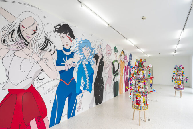 Eine moderne Kunstausstellung in einem hell erleuchteten Galerieraum. An der linken Wand ist ein großes, farbenfrohes Wandgemälde mit stilisierten, anime-inspirierten Figuren in kräftigen Farben zu sehen. Im Raum stehen zwei aufwendig dekorierte, bunte Skulpturen auf Holzstühlen, die aus Papier, Perlen und kleinen Objekten bestehen. Die Szenerie vermittelt eine verspielte und kulturell vielfältige Atmosphäre.