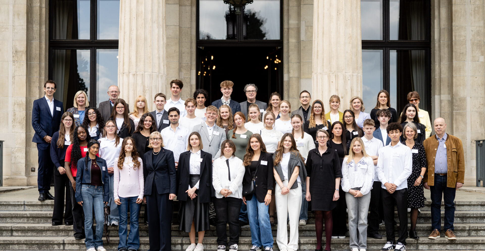 Gruppenfoto von etwa 45 Personen, bestehend aus Jugendlichen und Erwachsenen, die auf den Treppenstufen vor einem historischen Gebäude mit großen Säulen und Fenstern posieren. Die Personen sind formell bis leger gekleidet, viele lächeln in die Kamera. Das Bild vermittelt den Eindruck einer offiziellen Veranstaltung im Bildungskontext.