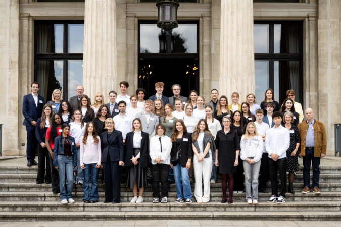 Gruppenfoto von etwa 45 Personen, bestehend aus Jugendlichen und Erwachsenen, die auf den Treppenstufen vor einem historischen Gebäude mit großen Säulen und Fenstern posieren. Die Personen sind formell bis leger gekleidet, viele lächeln in die Kamera. Das Bild vermittelt den Eindruck einer offiziellen Veranstaltung im Bildungskontext.