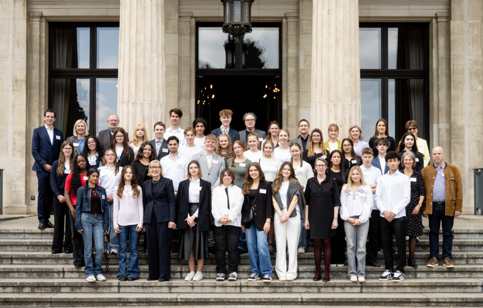Gruppenfoto von etwa 45 Personen, bestehend aus Jugendlichen und Erwachsenen, die auf den Treppenstufen vor einem historischen Gebäude mit großen Säulen und Fenstern posieren. Die Personen sind formell bis leger gekleidet, viele lächeln in die Kamera. Das Bild vermittelt den Eindruck einer offiziellen Veranstaltung im Bildungskontext.