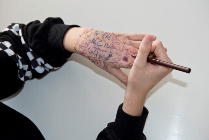 Ein paar Hände: Die rechte Hand schreibt mit einem Stift auf die linke Hand, die bereits voll geschrieben ist.