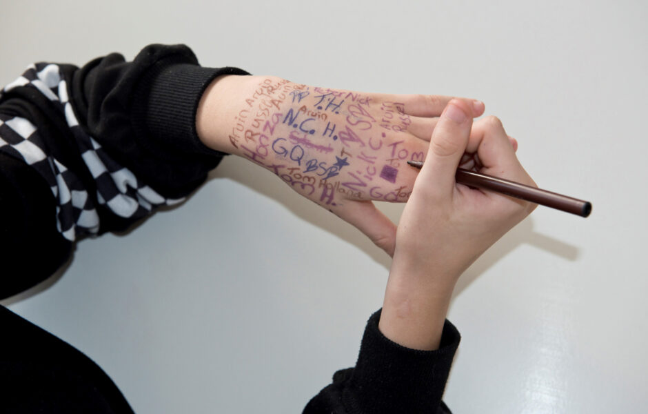 Ein paar Hände: Die rechte Hand schreibt mit einem Stift auf die linke Hand, die bereits voll geschrieben ist.