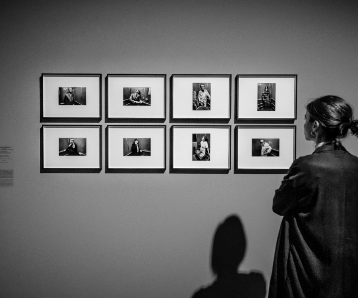 Woman standing in front of a wall with several framed portrait photographs arranged in two rows, looking at the images.