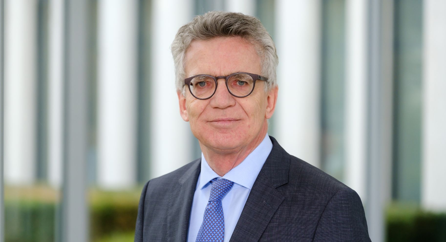 Dr. Thomas de Maizière, im dunklen Anzug mit blauer Krawatte als Portrait vor unscharfem Hintergrund.