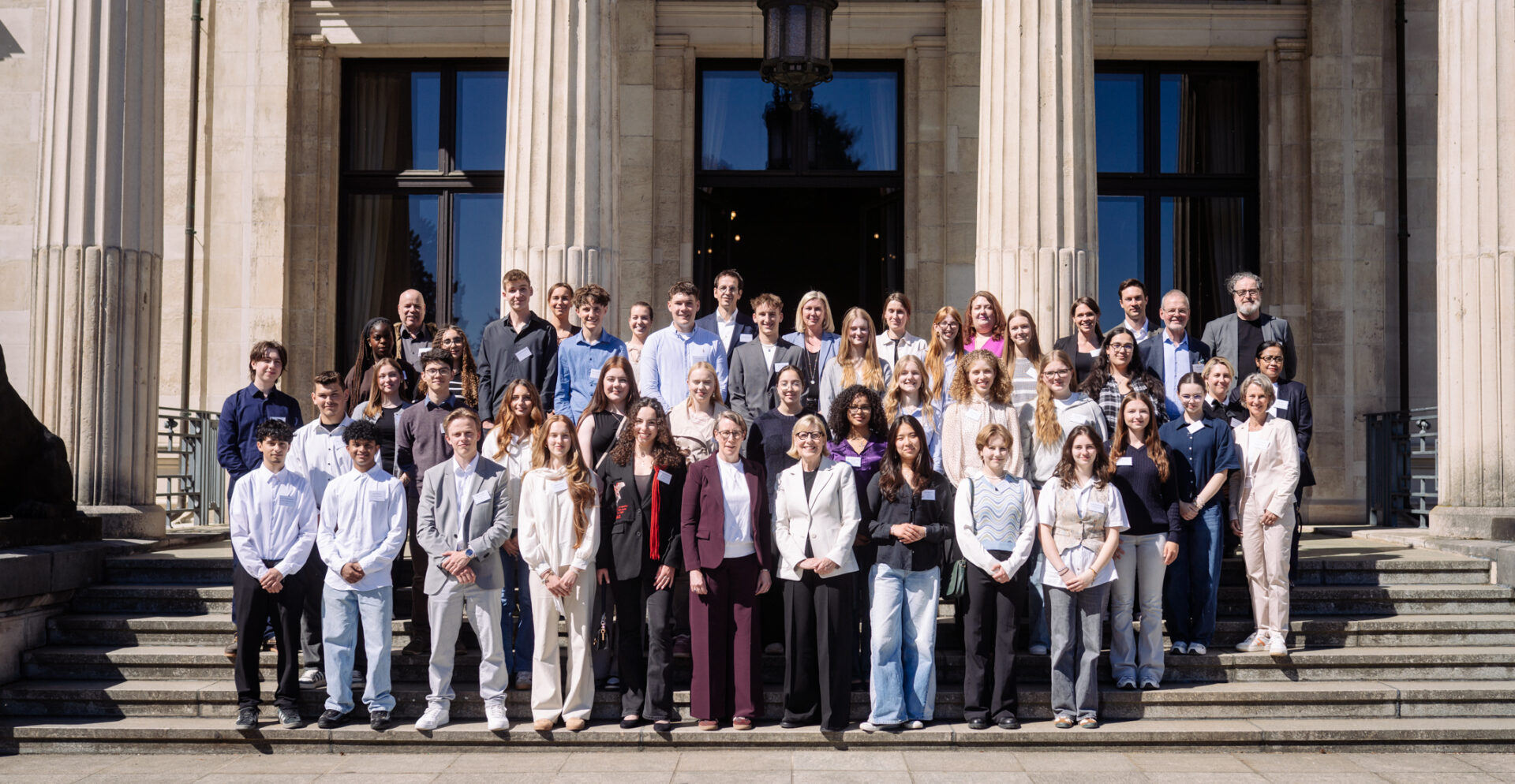 Eine Gruppe Schüler*innen mit ein paar Erwachsenen auf einer Treppe im Hintergrund ein Gebäude Säulen.