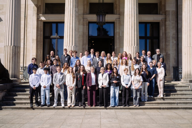 Eine Gruppe Schüler*innen mit ein paar Erwachsenen auf einer Treppe im Hintergrund ein Gebäude Säulen.