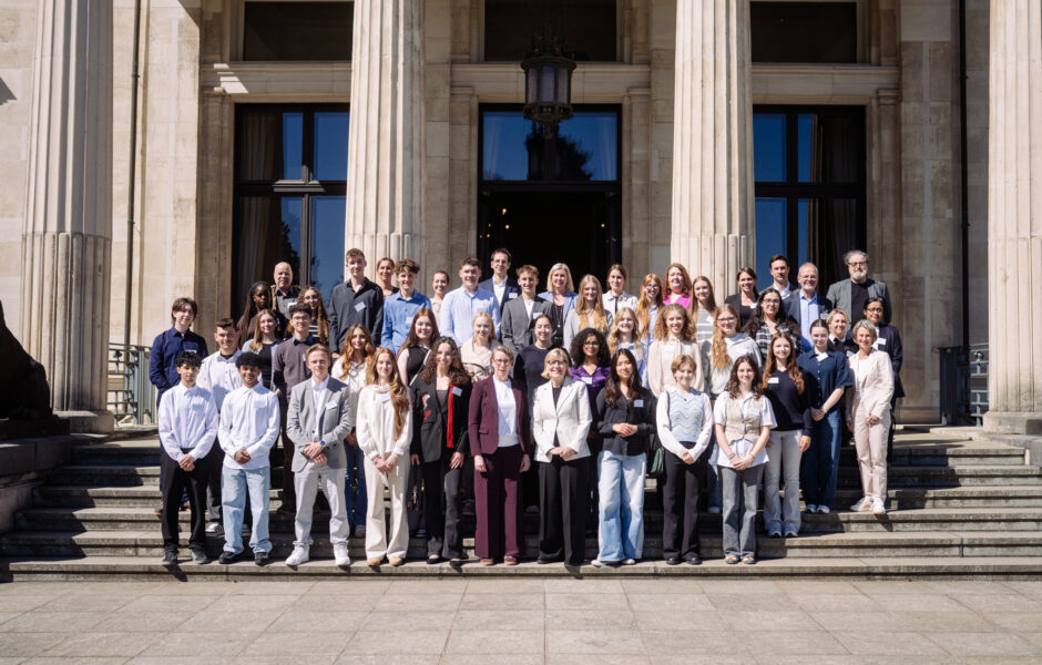 Eine Gruppe Schüler*innen mit ein paar Erwachsenen auf einer Treppe im Hintergrund ein Gebäude Säulen.