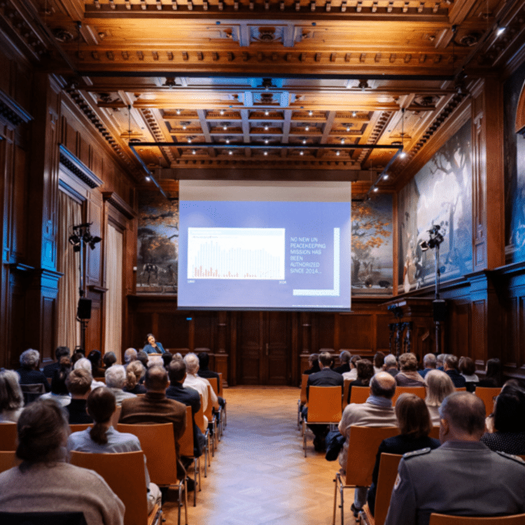 Mehrere Reihen von Besuchenden sitzen in einem holzvertäfelten Saal vor einer großen Projektionsleinwand.