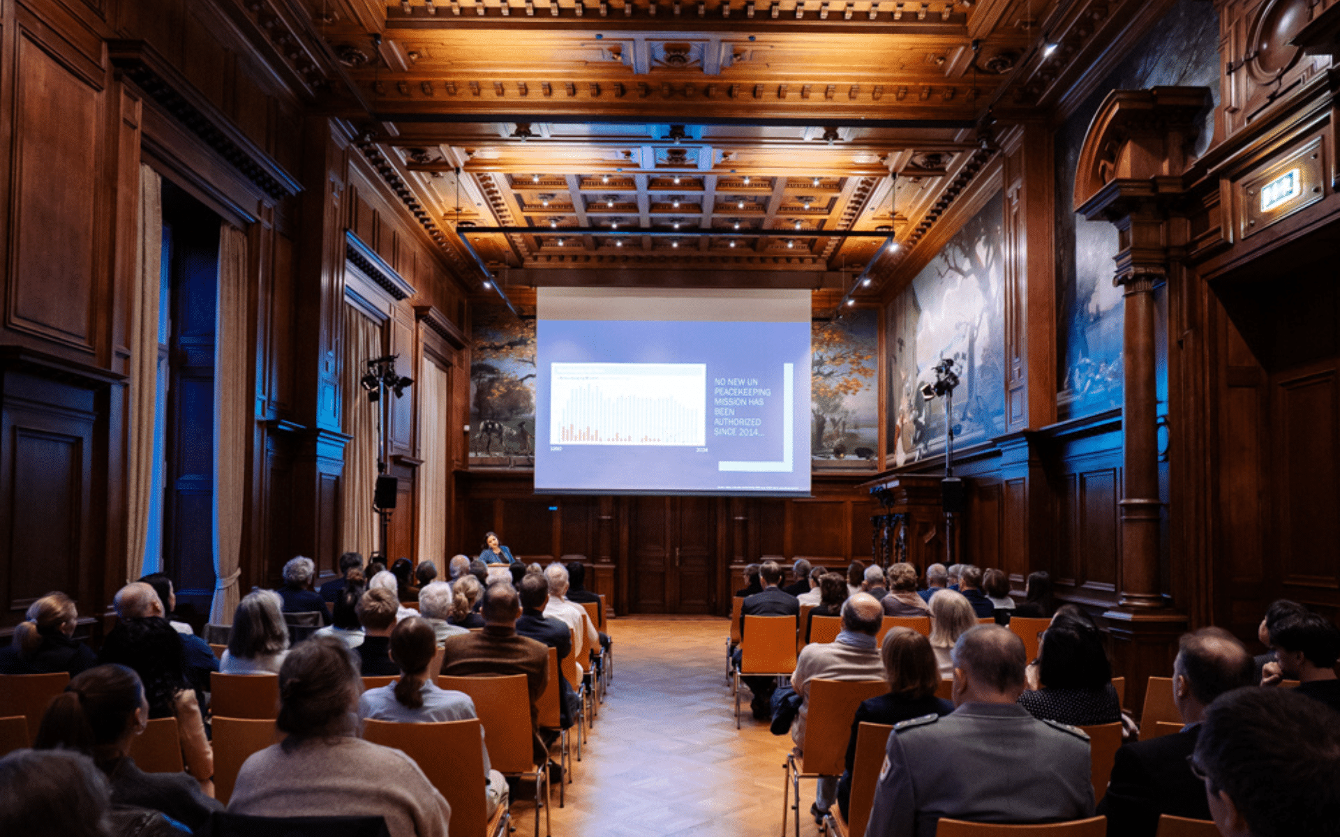 Mehrere Reihen von Besuchenden sitzen in einem holzvertäfelten Saal vor einer großen Projektionsleinwand.