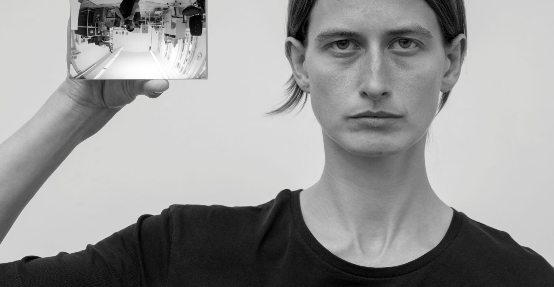 Eine Person im Portrait die einen Handspiegel neben ihrem Kopf in Richtung Kamera hält. Dort spiegelt sich der Raum und der Fotograf auf dem Kopf.