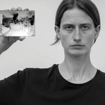 Eine Person im Portrait die einen Handspiegel neben ihrem Kopf in Richtung Kamera hält. Dort spiegelt sich der Raum und der Fotograf auf dem Kopf.