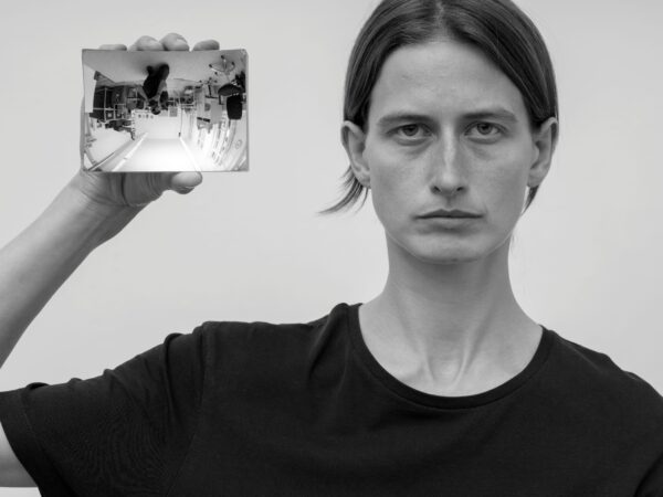 Eine Person im Portrait die einen Handspiegel neben ihrem Kopf in Richtung Kamera hält. Dort spiegelt sich der Raum und der Fotograf auf dem Kopf.