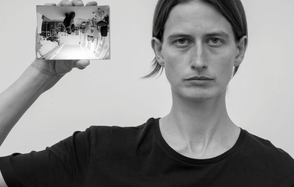 Eine Person im Portrait die einen Handspiegel neben ihrem Kopf in Richtung Kamera hält. Dort spiegelt sich der Raum und der Fotograf auf dem Kopf.