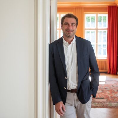 Ein Portrait von Prof. Dr. William Burke-White, der an eien Wand lehnt. Im Hintergrund eröffnet sich ein Raum.