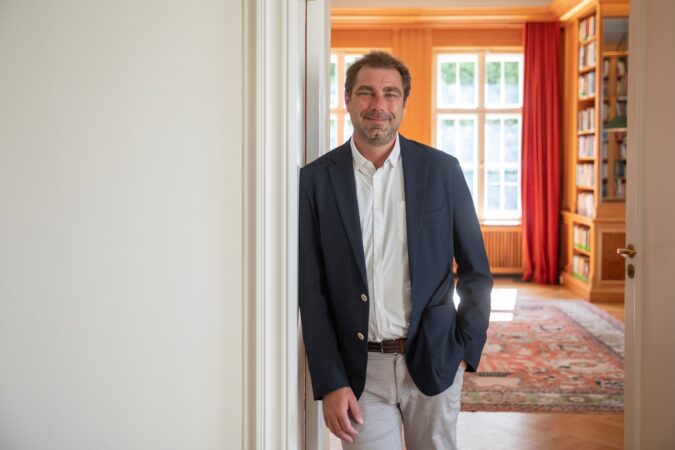 Ein Portrait von Prof. Dr. William Burke-White, der an eien Wand lehnt. Im Hintergrund eröffnet sich ein Raum.