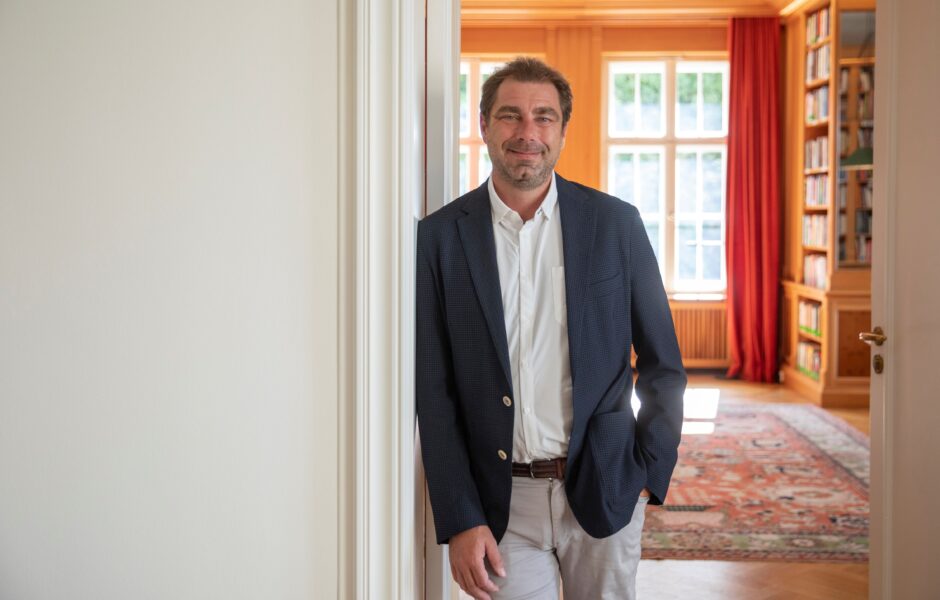 Ein Portrait von Prof. Dr. William Burke-White, der an eien Wand lehnt. Im Hintergrund eröffnet sich ein Raum.