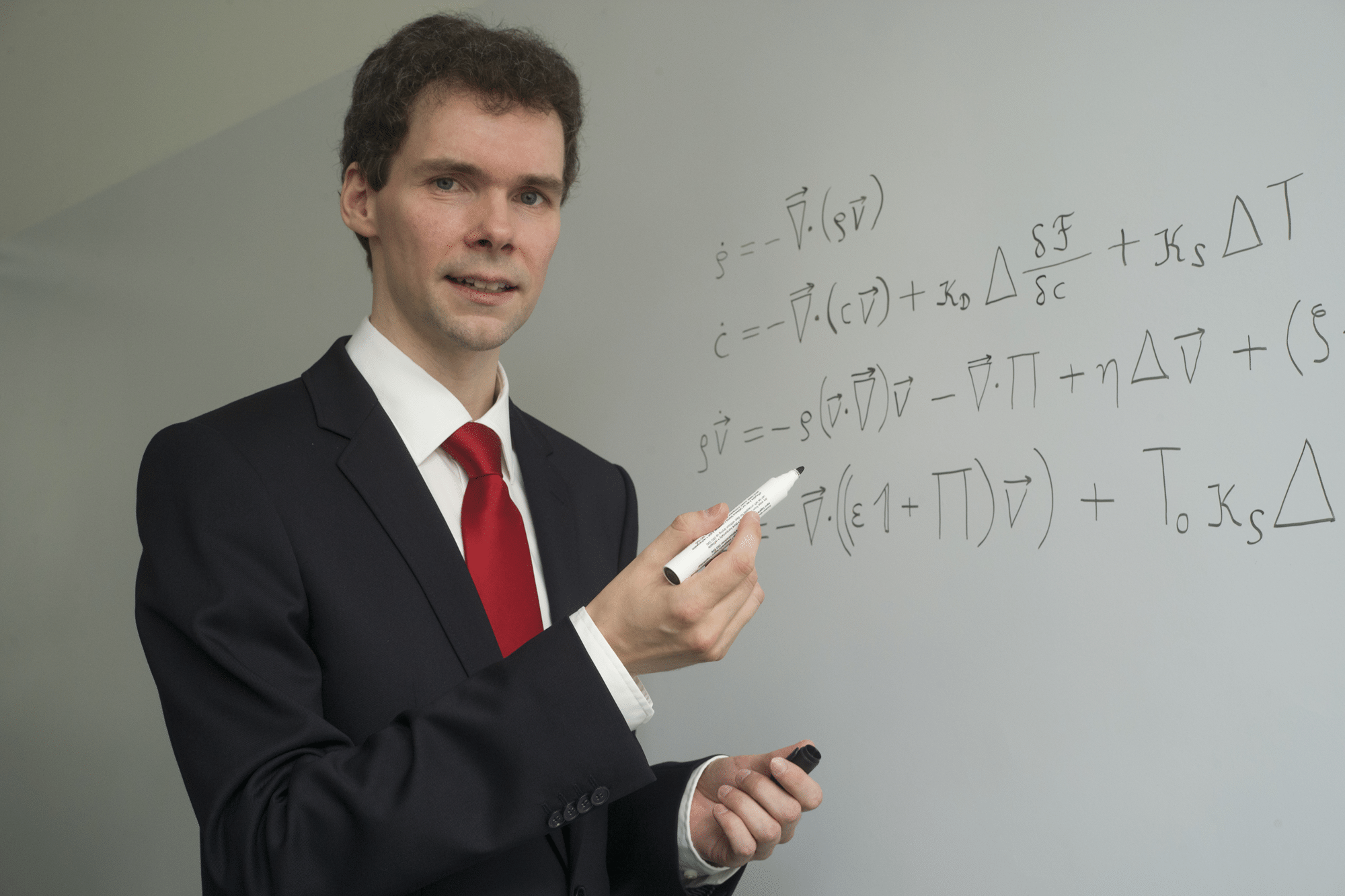 Eine Person im Anzug hält einen Stift vor eine beschriebene Tafel mit mathematischen Formeln und Symbolen.